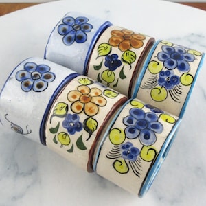 May include: Six ceramic napkin rings, each hand-painted with floral patterns in blue, orange, and yellow on a cream base. The rings are cylindrical and displayed on a white marble surface. The design is reminiscent of traditional pottery.