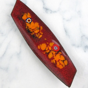 May include: A rectangular, red enamel barrette with orange and yellow floral designs. The barrette features two small, colorful floral accents and a speckled texture. The accessory is likely for hair styling.
