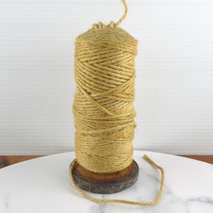 May include: A spool of natural-colored twine, tightly wound, stands on a wooden base. The twine is a light brown color, and a length of it extends from the top and side. The spool is set on a white marble-like surface.