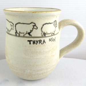 May include: Cream-colored ceramic mug with a curved handle. The mug features a black line drawing of three sheep and the text "THYRA NSW" below the sheep. The mug has a slightly textured surface and a light brown rim.