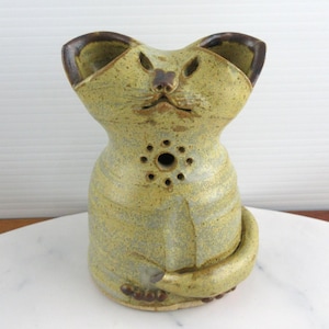 May include: A ceramic cat-shaped vessel with a speckled yellow-green glaze. The figure features pointed ears, a small nose, and a circular cutout on the chest. A curved tail wraps around the base, and the rim is accented with a dark brown finish.