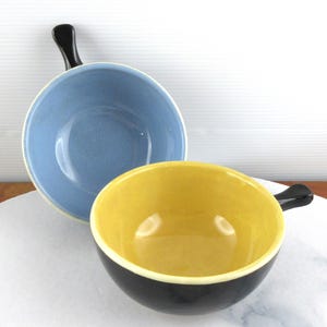 May include: Two ceramic bowls with handles. One is light blue, the other is yellow with a black exterior. Both have a black handle. The bowls are on a white marble surface.