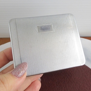 May include: A silver rectangular compact with a textured surface and a small rectangular accent. The compact is held in a hand, resting on a brown leather surface.