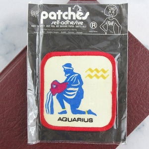 May include: A self-adhesive Aquarius patch in a clear plastic package. The patch features a blue figure pouring water from a red vessel, with the word "AQUARIUS" below. The patch has a red border and yellow wavy lines.