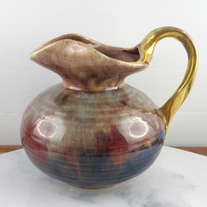 May include: A small, round ceramic pitcher with a gold handle. The pitcher has a brown, purple, and green glaze with a glossy finish.