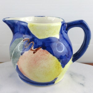 May include: A ceramic pitcher with a blue background and a colorful fruit design. The pitcher has a handle and a spout. The fruit design includes yellow, pink, and green elements. The interior is white.