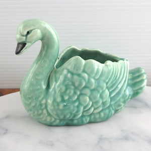 May include: A light green ceramic swan planter featuring a glossy finish and textured feather details on the body. The swan has a long, curved neck and a dark beak, with an open cavity on its back for holding small items or plants.