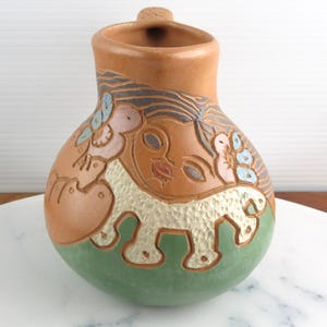 May include: A terracotta pitcher featuring a rounded body with a green base and a carved relief design. The surface displays a stylised face with flowing hair, floral accents in blue and pink, and a textured white animal figure at the bottom.