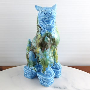 May include: A ceramic foo dog figurine featuring a glossy glaze in shades of light blue, dark green, and brown. The mythical creature is depicted in a seated position on a rectangular base, with detailed facial features and a textured mane.