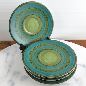 Op de afbeelding: Set van vier keramische borden met concentrische ringen in turquoise, groen en donkergroen. De borden zijn gestapeld en gepresenteerd op een wit marmeren oppervlak, met twee borden op een zwarte standaard. Retro esthetiek.