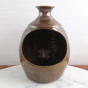 May include: A brown ceramic salt cellar with a glossy finish and a wide, circular front opening. The vessel features a narrow neck at the top and the word Salt embossed in a decorative script font along the lower front section of the body.
