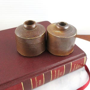 Puede incluir: Dos pequeños tinteros de cerámica marrón, de forma cilíndrica, con una pequeña abertura en la parte superior. Se encuentran sobre un libro granate con letras doradas y un marcapáginas de cinta roja. Aspecto rústico y antiguo.