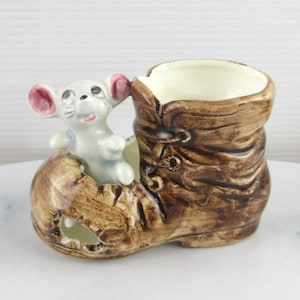 May include: A ceramic figurine of a brown boot with a small grey mouse peeking out of the top. The boot has a white interior and brown laces.