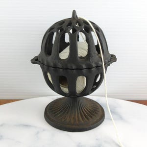 Puede incluir: Un portacuerdas de hierro fundido negro con un diseño de jaula esférica calada y una base de pedestal. El soporte contiene un carrete de cuerda blanca, con un solo hilo que se extiende a través de la abertura superior de la estructura.