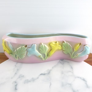 Puede incluir: Una jardinera de cerámica rosa con interior azul claro y forma curva. La jardinera está decorada con hojas en relieve en verde, amarillo y azul, con detalles dorados. La jardinera está sobre una superficie de madera.