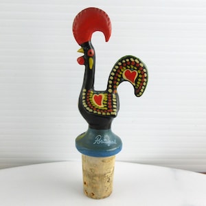 May include: A decorative bottle stopper with a rooster design. The rooster has a red comb, black body, and a tail with red heart accents. The base is blue with the word "Portugal" written on it.
