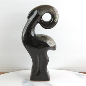 May include: A dark brown ceramic sculpture with a distinctive, curved form. The sculpture has a glossy finish with a lighter, speckled pattern near the top. The base is rectangular, and the top has a circular opening.
