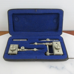 May include: A vintage drafting instrument set featuring two silver-toned metal compass tools with adjustable knobs and a small cylindrical attachment, all housed within a black rectangular carrying case lined with vibrant blue felt interior.