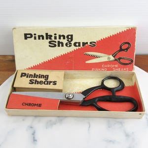 Puede incluir: Unas tijeras de zig-zag cromadas con mangos negros dentro de una caja de cartón rectangular. La tapa muestra el texto Pinking Shears y un gráfico de las tijeras, el interior está forrado con papel de puntos rojos y blancos.