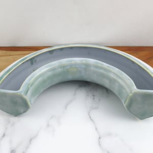 Puede incluir: Un macetero de cerámica azul claro con forma de media luna. El macetero tiene un acabado esmaltado y un tono amarillo sutil.