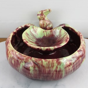 Könnte beinhalten: Eine dekorative Keramikschale mit zweistufigem Design. Die Schale ist überwiegend bordeauxrot mit grünen und gelben Akzenten. Eine kleine Hasenfigur sitzt auf der oberen Ebene. Die Schale dient wahrscheinlich dekorativen Zwecken.