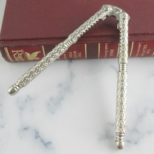 Puede incluir: Cascanueces plateado con un diseño decorativo y estampado. El cascanueces está abierto, revelando su mecanismo interior. Descansa sobre una superficie de mármol con un libro granate en el fondo.