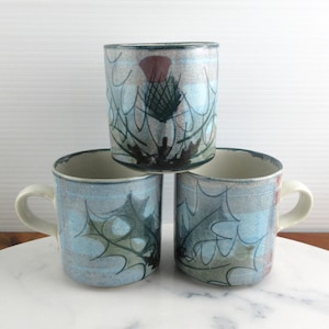 May include: Three ceramic mugs stacked in a pyramid formation. Each mug features a light blue and white plaid background with dark green thistle and holly leaf illustrations. The mugs have a cylindrical shape with a single handle on the side.
