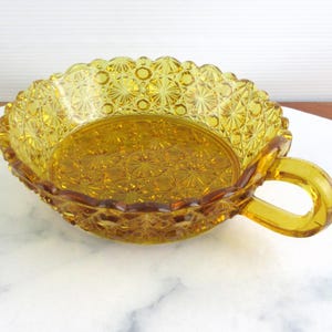 May include: Amber glass serving dish with a handle. The dish features a starburst pattern on the base and a scalloped rim. The handle is a loop shape. A vintage piece, likely from the mid-20th century.