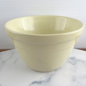 May include: A pale yellow ceramic mixing bowl with a smooth, glossy finish. The bowl features a wide, open top with a distinct horizontal ridge encircling the upper exterior, tapering down to a rounded base with a consistent light colour.