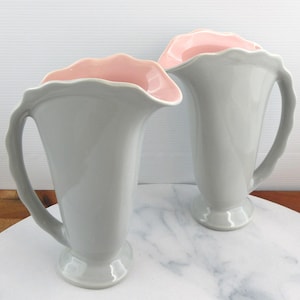 May include: Two grey ceramic jugs with a pink interior and scalloped rims. Each jug has a curved handle and a pedestal base. The jugs are displayed on a marble-patterned surface.