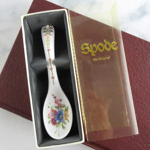 Peut inclure: Une cuillère en porcelaine blanche avec un motif floral, présentée dans une boîte. La cuillère présente un motif détaillé de fleurs roses, bleues et jaunes. La boîte a un couvercle en plastique transparent et le texte "Spode The Original".