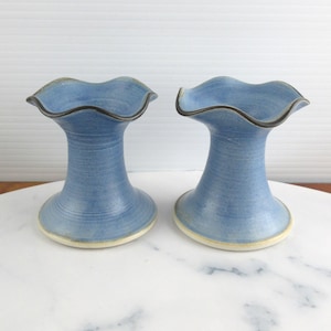 May include: A pair of blue ceramic candle holders with a flared, wavy rim and a dark brown edge. The holders feature a smooth, tapered body that widens at the base, showcasing a subtle textured finish in shades of light and medium blue.