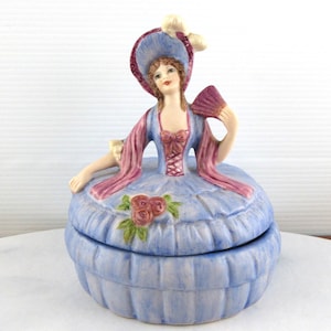 May include: A blue and white porcelain trinket box with a figurine of a woman in a blue dress and a pink shawl holding a fan. The box is decorated with pink roses.