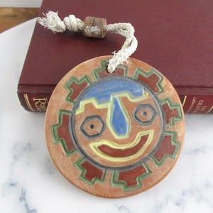 May include: A round ceramic sun face pendant with a rope. The pendant features a smiling face with blue, yellow, and red accents. The sun face is surrounded by a green and red border. The pendant is brown.