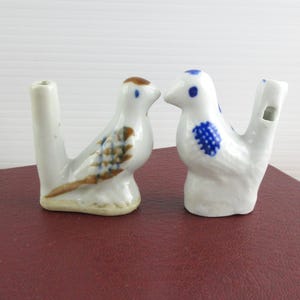 May include: Two ceramic bird whistles. One bird has brown and blue wing details, the other has blue accents. Both are white with a small opening for the whistle. The birds are perched on a dark red surface.