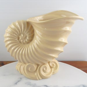 May include: A cream-coloured ceramic vase shaped like a nautilus shell. The vase features a spiralled, ribbed exterior texture and rests on a base decorated with sculpted, swirling wave-like patterns. The finish is smooth and glossy throughout.