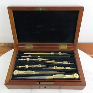 May include: A rectangular wooden box with a hinged lid, containing a set of six brass and metal drafting instruments arranged in a dark blue velvet-lined interior. The tools include various compasses and dividers with metallic finishes.