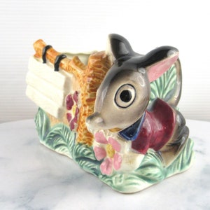 May include: A ceramic planter shaped like a rabbit holding a sign. The rabbit is grey with black eyes and a red and blue waistcoat. The planter is decorated with green leaves, pink flowers, and a brown branch.