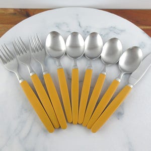 Puede incluir: Un juego de cubiertos de acero inoxidable con mangos amarillos, que incluye tenedores, cucharas y un cuchillo. El juego está dispuesto sobre una superficie de mármol blanco. Utensilios para comer.