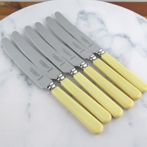 May include: A collection of seven butter knives. Each knife has a stainless steel blade and a pale yellow handle. The knives are displayed on a white marble surface. Text is printed on each blade.
