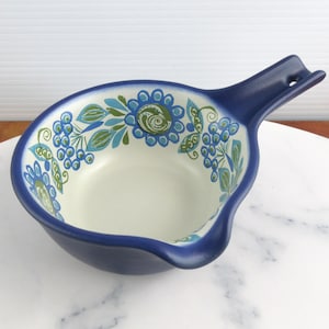 May include: A deep blue ceramic bowl with a handle and a pouring spout. The interior features a cream-colored base decorated with a repeating pattern of blue and green stylized flowers, leaves, and small berry clusters around the inner rim.