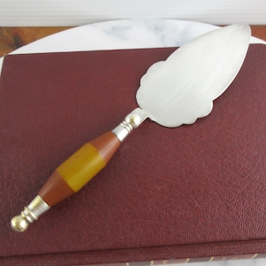 May include: A silver cake server with a scalloped edge blade and a handle featuring a brown and yellow design. The server is resting on a burgundy book.