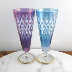 May include: Two iridescent champagne flutes, one purple, one blue, with a diamond pattern and gold rims. The flutes are on a white surface.
