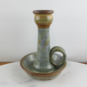 Puede incluir: Un candelabro de cerámica con un esmalte moteado en tonos verde salvia, azul y marrón. La pieza tiene una base circular ancha, una columna central con la parte superior acampanada y una pequeña asa redondeada unida al lado de la base.