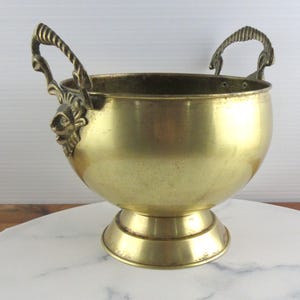 May include: A polished brass bowl with a rounded body and a pedestal base. The bowl features two ornate handles, each decorated with a sculpted lion head motif at the base and a ribbed, curved design extending upward from the rim of the vessel.