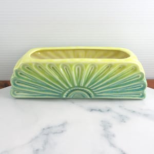 May include: A rectangular ceramic planter featuring a vibrant sunburst pattern on the front. The design transitions from a deep teal at the base to a bright yellow at the top rim, with raised, radiating lines creating a textured, fan-like effect.