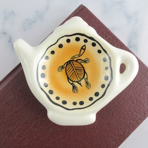 May include: Cream-colored ceramic tea bag holder in a teapot shape. The center features an orange circle with a black turtle design. The rim is decorated with black dots. The holder rests on a dark brown surface.