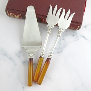 May include: A silver cake server and two silver forks with amber-coloured handles. The serving utensils are arranged on a white marble surface next to a closed, maroon-coloured book.