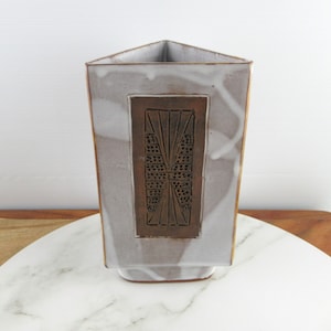 Könnte beinhalten: Eine weiße, dreieckige Keramikvase mit einem braunen geometrischen Muster. Die Vase hat eine strukturierte Oberfläche und einen quadratischen Boden.