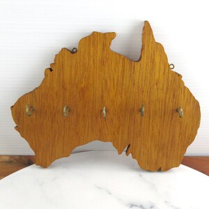 May include: A wall-mounted key holder crafted from light brown wood, shaped in the silhouette of the Australian continent. The wooden piece features five small, evenly spaced brass-colored metal hooks attached to the front for hanging keys.